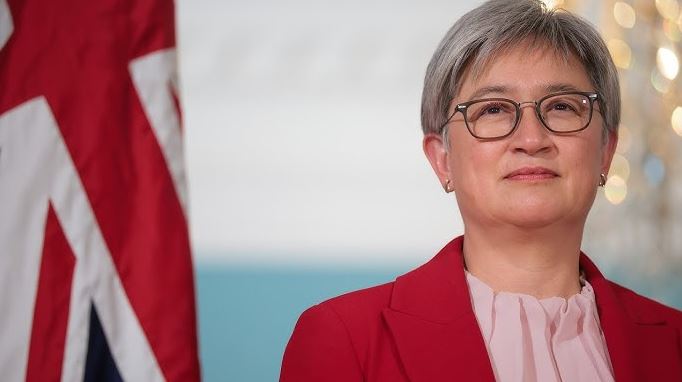 Portrait of a Penny Wong in a red blazer and glasses standing in front of an Australian flag, in a formal political setting