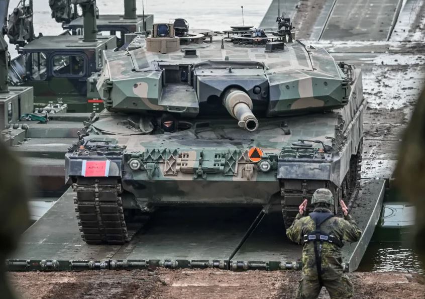 Armored military tank crossing a temporary bridge during a military exercise, with soldiers guiding the vehicle in a muddy combat training environment.