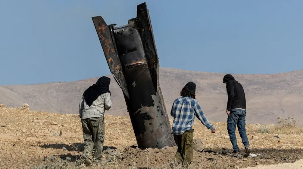 Civilians inspecting a damaged missile or rocket fragment in a desert area following military strikes in the Middle East conflict zone.
