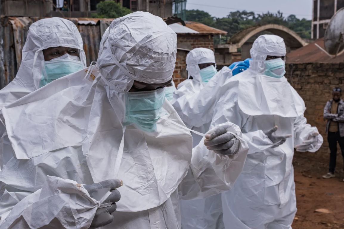 Healthcare workers wearing full-body protective suits, masks, and gloves prepare Ebola safety equipment in an outdoor area during an outbreak response operation in Africa.