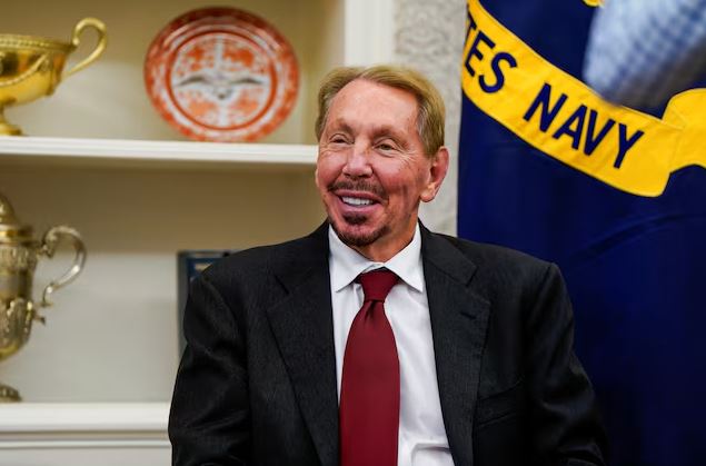 Larry Ellison smiling during an interview in a formal setting with trophies and a U.S. Navy flag in the background.